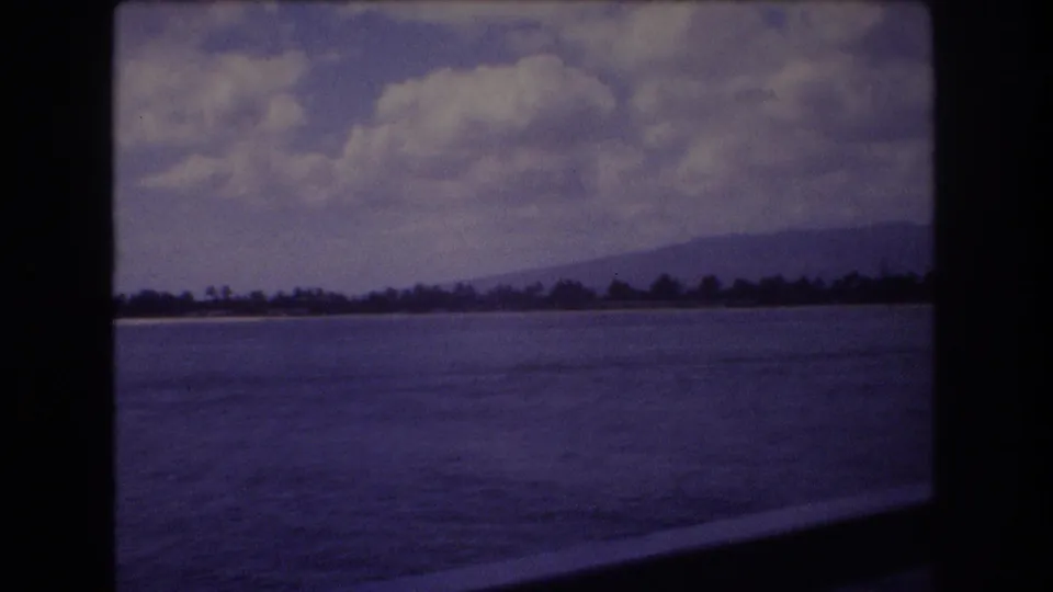 1977 Honolulu 8mm frame showing blue water, a low tree-lined shore, mountains in the distance, and large clouds across the sky.