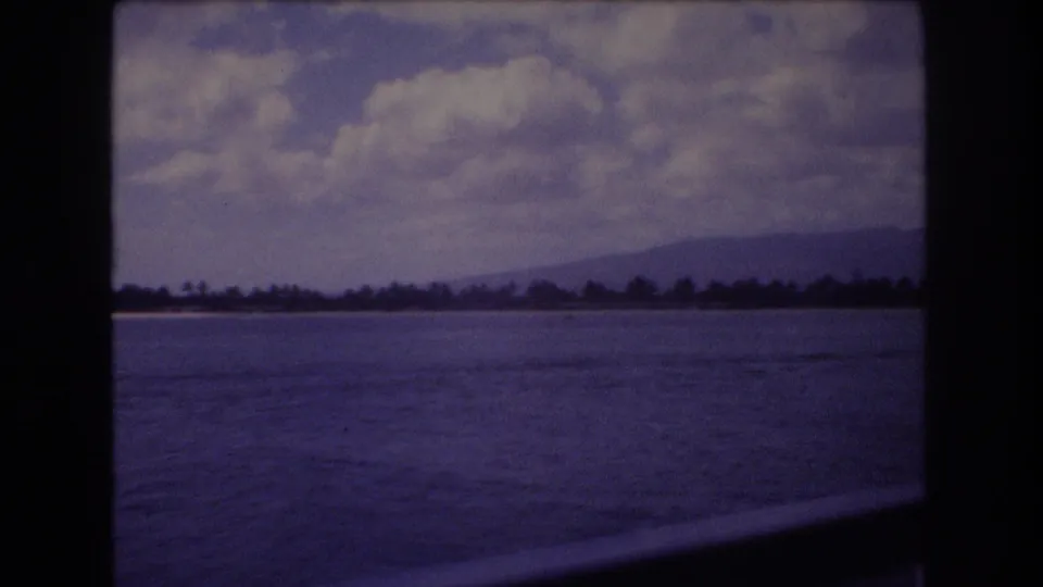 1977 Honolulu coastal home-movie frame with water in the foreground, a distant shore and mountain ridge, cloud cover overhead, and visible dark 8mm frame edges.