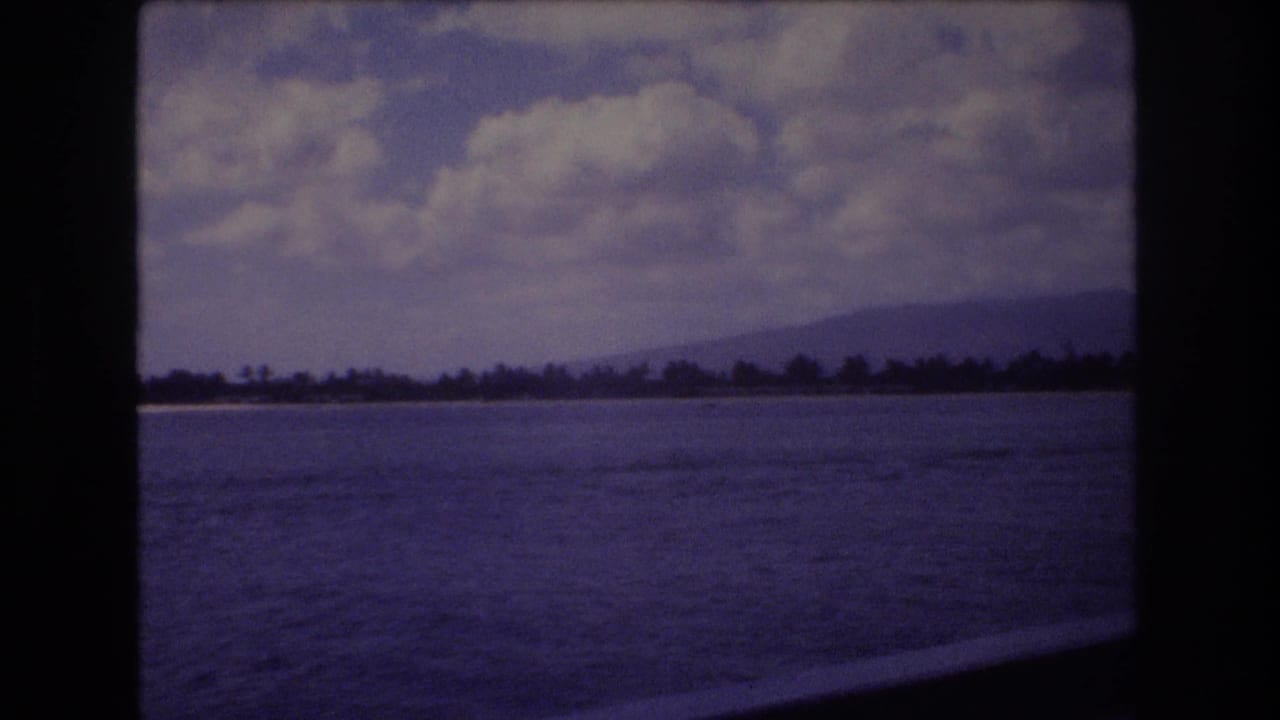 1977 8mm home-movie frame from Honolulu, Hawaii, showing blue ocean water from a moving vessel with a distant tree-lined shoreline, low mountains, and white clouds.