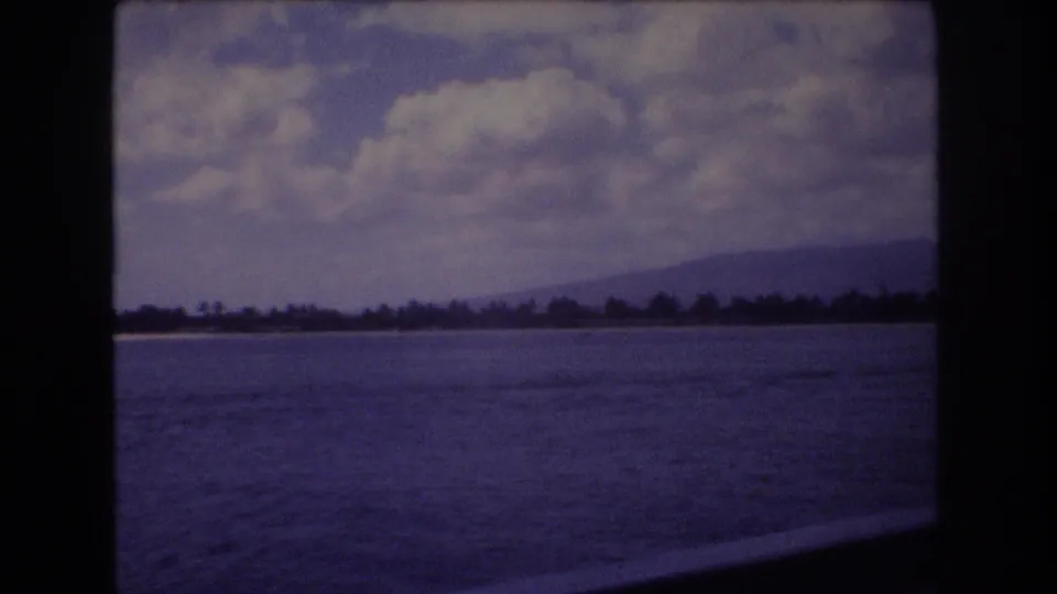 1977 8mm home-movie frame from Honolulu, Hawaii, showing blue ocean water from a moving vessel with a distant tree-lined shoreline, low mountains, and white clouds.