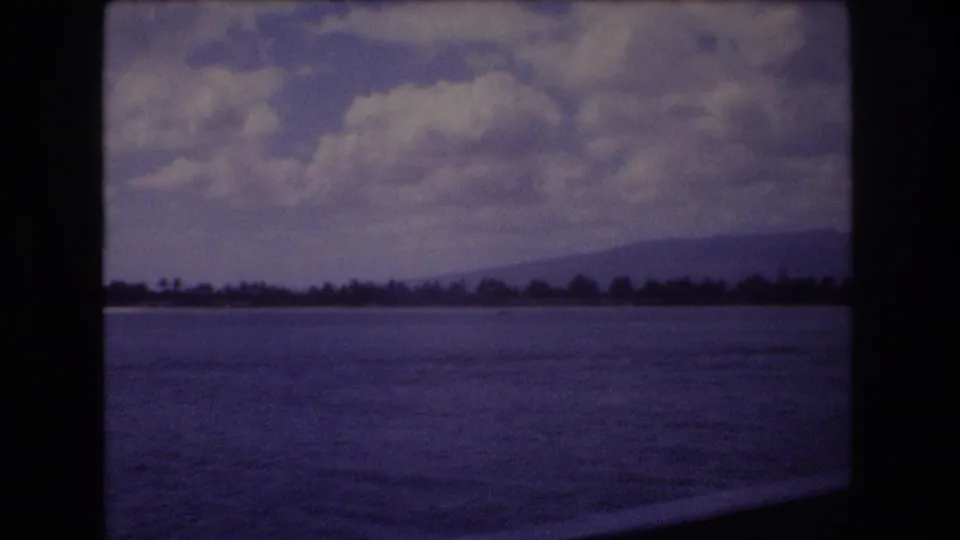 1977 8mm home-movie view near Honolulu, Hawaii, looking across rippled ocean water toward a low shoreline, dark trees, mountains, and a cloud-filled sky.