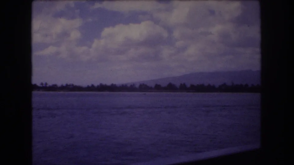 1977 8mm home-movie frame from Honolulu, Hawaii, with blue water in the foreground, a distant coastal tree line, low mountains, and large white clouds overhead.