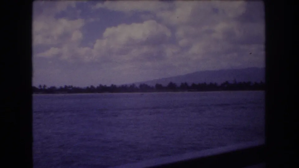 1977 8mm home-movie view near Honolulu, Hawaii, showing ocean surface, a far tree-lined shoreline, low mountains, and soft clouds in a purple-tinted film frame.