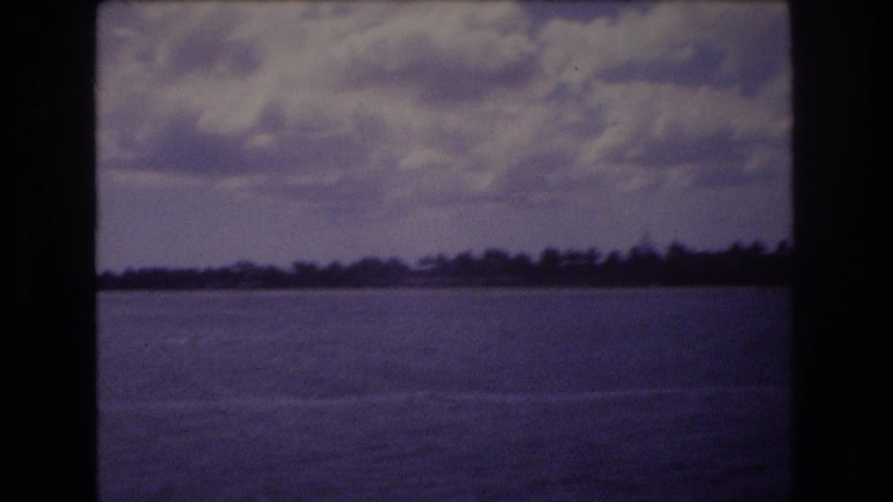 1977 Honolulu 8mm frame showing calm coastal water, a low distant shoreline, and cloudy sky with dark film borders.
