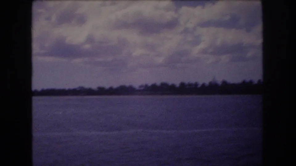 1977 Honolulu 8mm frame showing calm coastal water, a low distant shoreline, and cloudy sky with dark film borders.