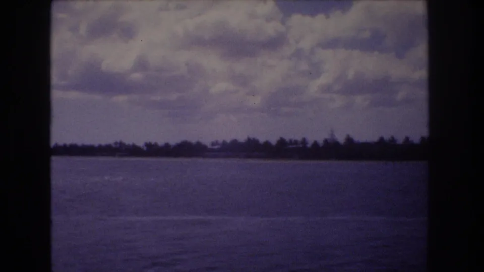 1977 Honolulu home-movie frame with open water in the foreground and a low palm-lined shoreline under heavy clouds.
