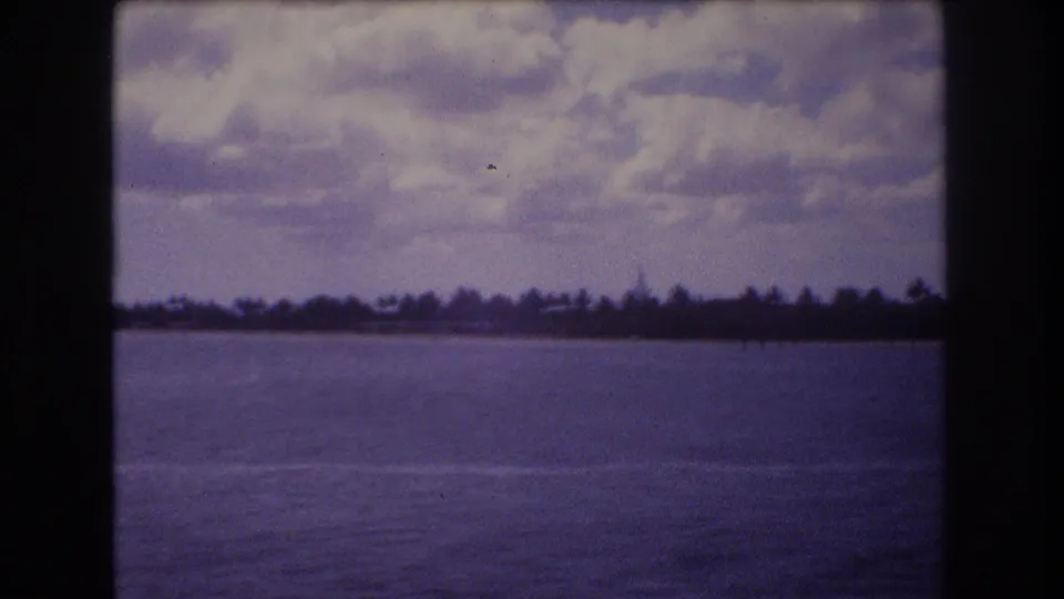 1977 Honolulu 8mm frame showing blue-purple water, a narrow shoreline strip, dark trees, and textured cloud cover.