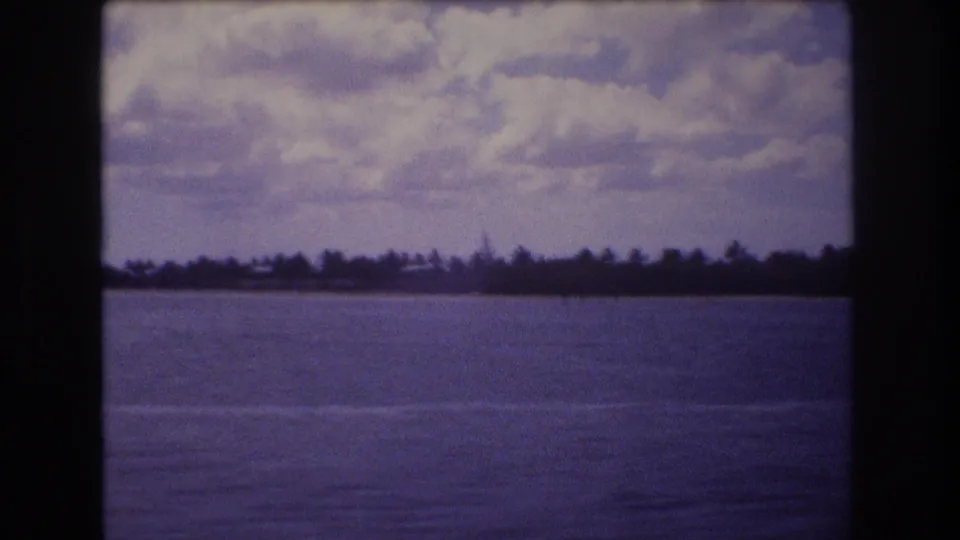 1977 Honolulu travel frame with calm water, a faint shore edge, silhouetted trees, and a cloudy sky in grainy 8mm color.