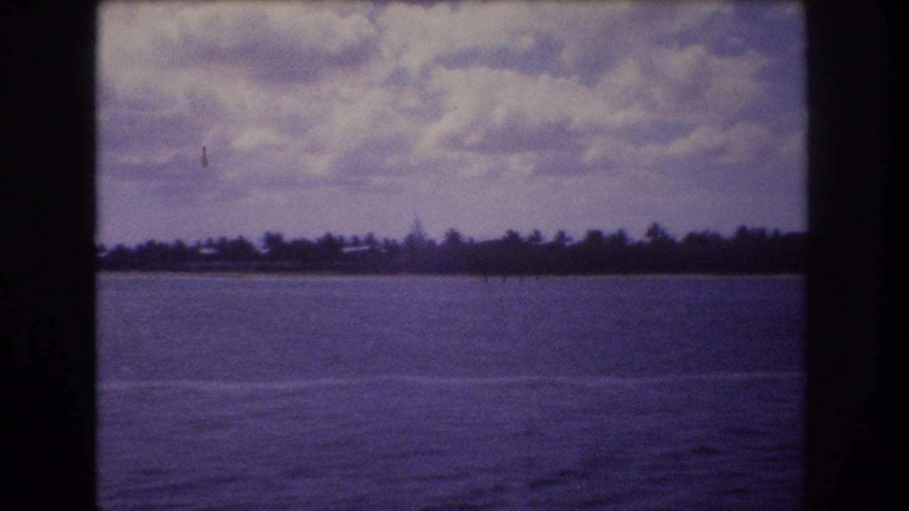 1977 8mm Honolulu-area vacation view from a moving boat, showing open water, a low palm-lined shore, and a cloudy sky.