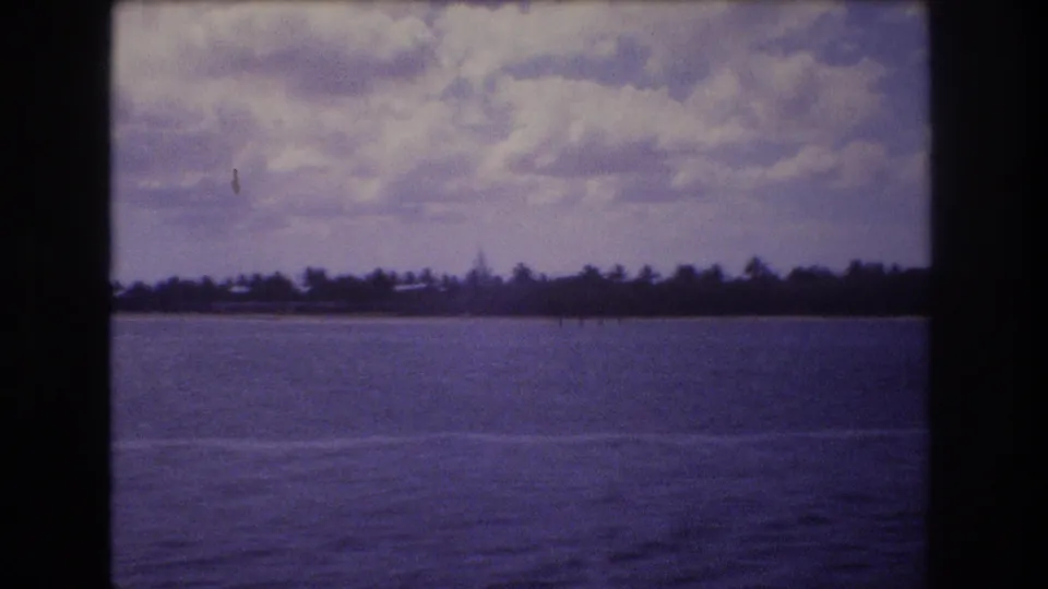 1977 8mm Honolulu-area vacation view from a moving boat, showing open water, a low palm-lined shore, and a cloudy sky.
