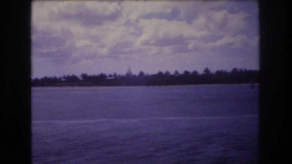 A second 1977 home-movie view across the water toward a dark tree-lined shoreline, filmed from a moving boat in the Honolulu area.