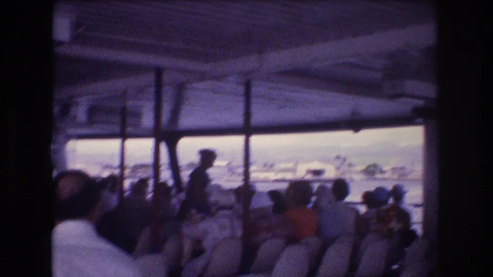Inside a passenger boat or ferry, seated adults face the water while a standing figure appears near the windows, with harbor scenery visible beyond.