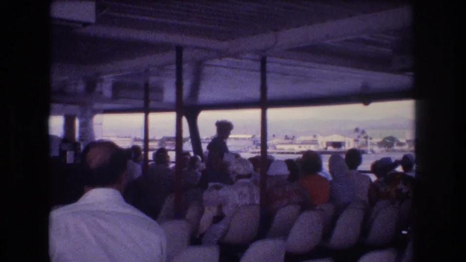 1977 Honolulu-area home-movie frame showing passengers seated beneath a shaded boat canopy, with water, boats, and low waterfront structures visible through the openings.