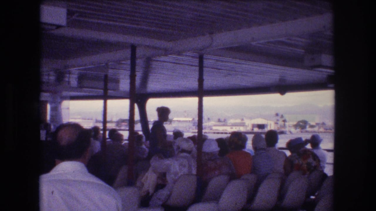 1977 Honolulu home-movie frame showing seated passengers on an open-sided vessel, with roof supports, water, and distant shoreline buildings visible outside.