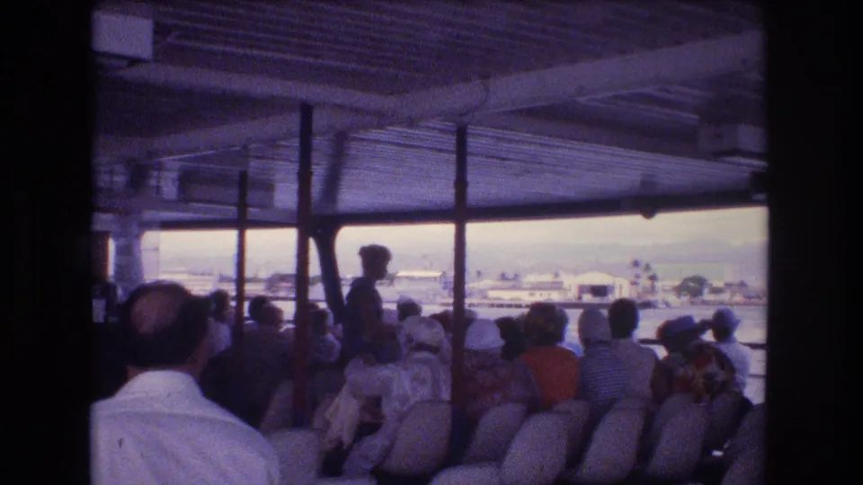 1977 Honolulu home-movie frame showing seated passengers on an open-sided vessel, with roof supports, water, and distant shoreline buildings visible outside.