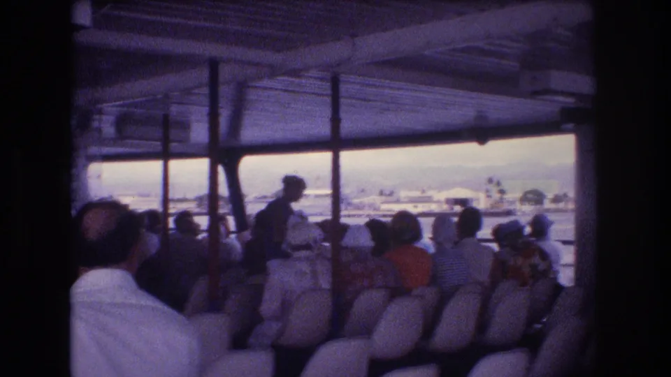 1977 Honolulu 8mm frame from aboard an open-sided passenger vessel, showing travelers seated beneath a roof and looking toward water and shoreline buildings.