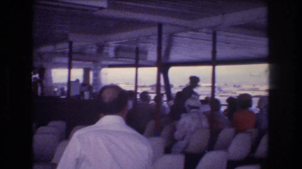 Final 1977 Honolulu frame showing the open-sided vessel interior with seated passengers, a standing figure near the aisle, and water with shoreline beyond.