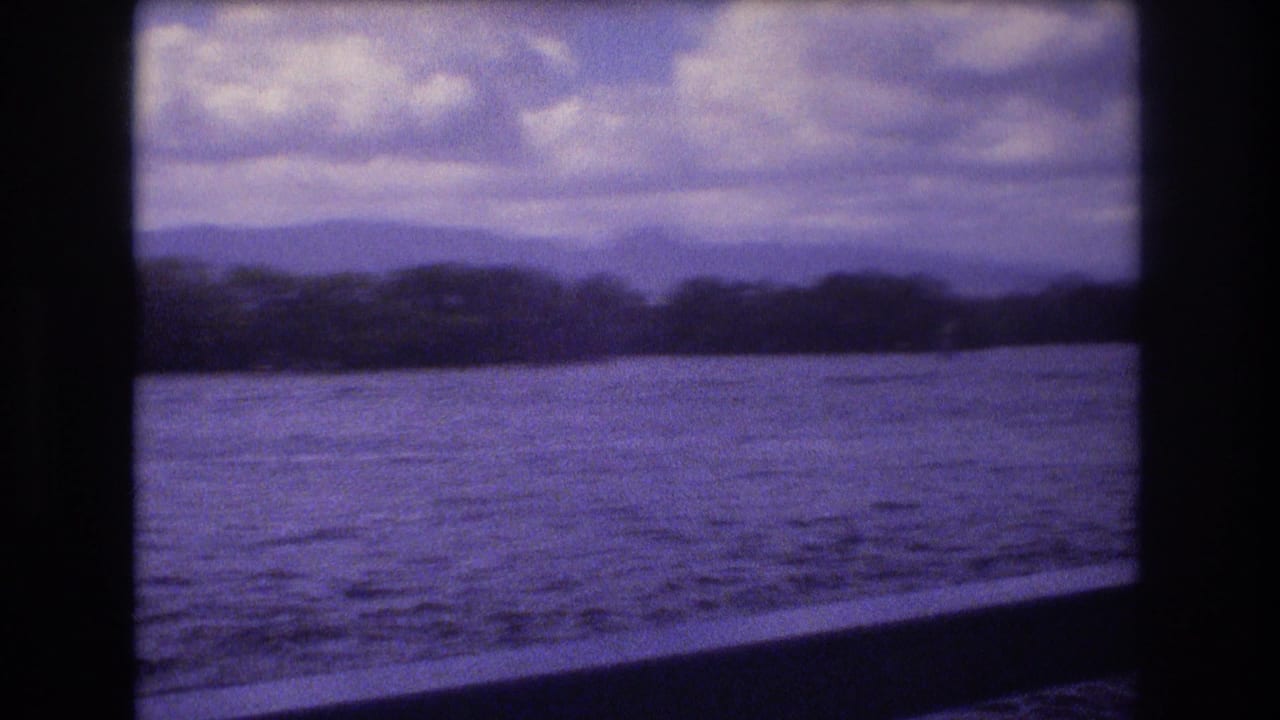 1977 Honolulu-area 8mm home-movie frame showing a boat-window view of rippled sea, low shoreline trees, distant mountains, and a cloudy sky with a purple film cast.