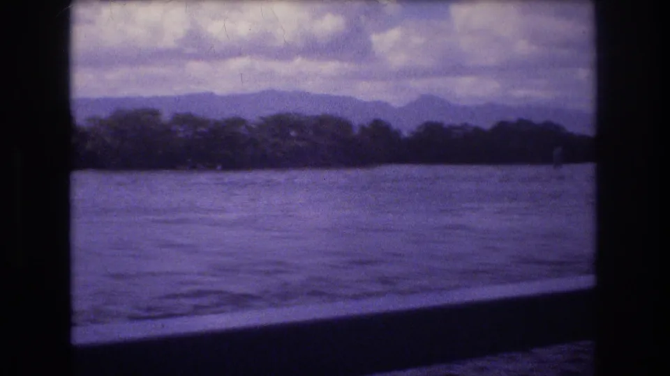 1977 Honolulu-area 8mm frame looking from a boat through dark framing at calm water, a distant wooded shoreline, mountain silhouettes, and heavy cloud cover.