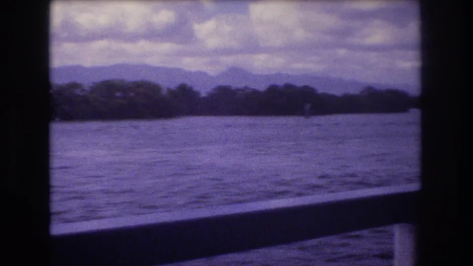 1977 Honolulu-area home-movie frame with a purple-tinted boat-window view over textured water toward trees, soft mountain outlines, and an overcast sky.
