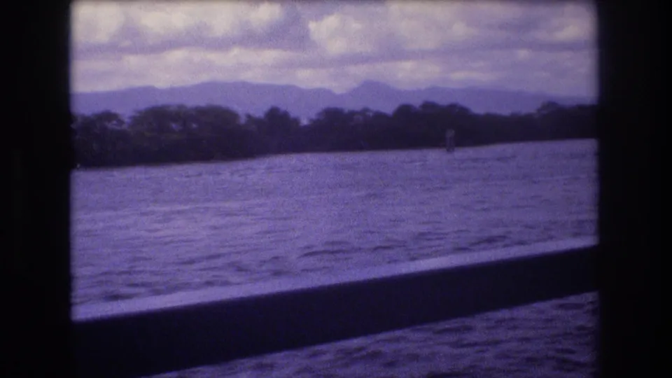 1977 Honolulu-area 8mm frame showing rippled water, a tree-lined shore, distant blue-purple mountains, and cloudy sky through a dark boat-window opening.