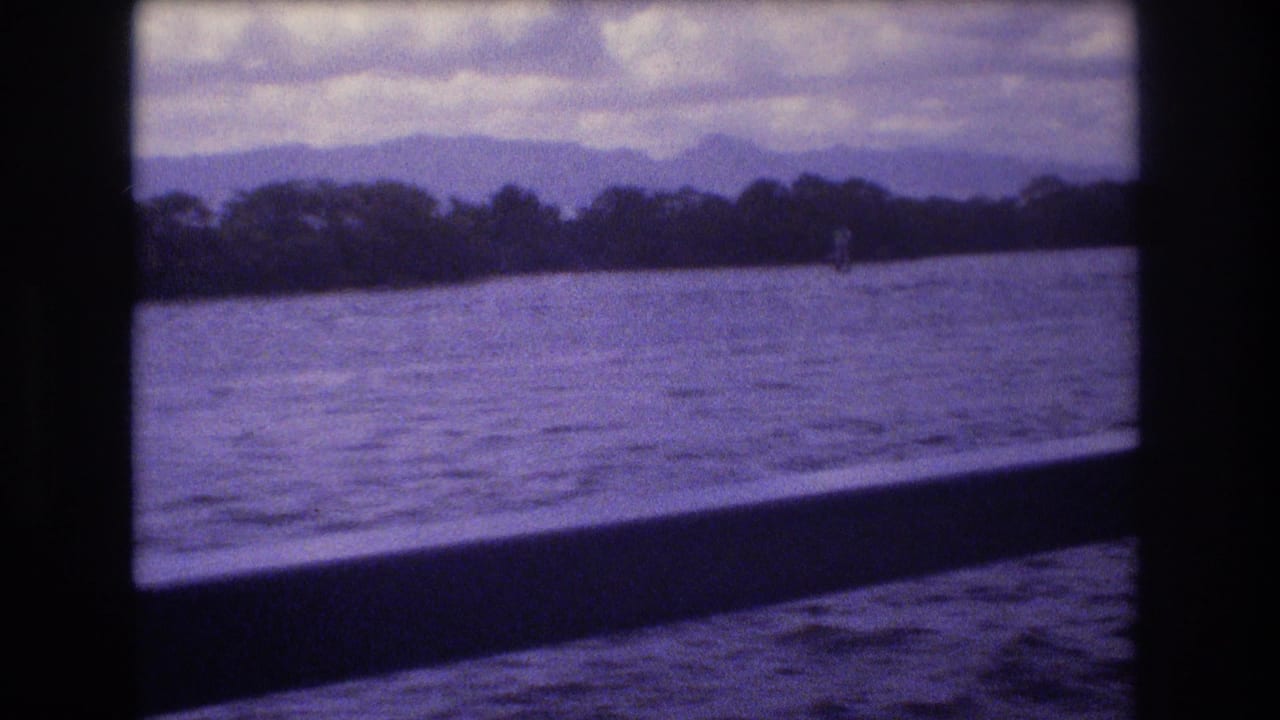 1977 Honolulu ferry view showing choppy harbor water, a dark window frame, distant shoreline trees, and mountains under a cloudy sky.
