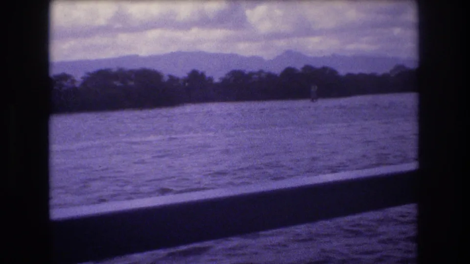 A Honolulu ferry window frames open water, a low tree-lined shore, and distant mountains with the soft purple-blue cast of aged 8mm film.