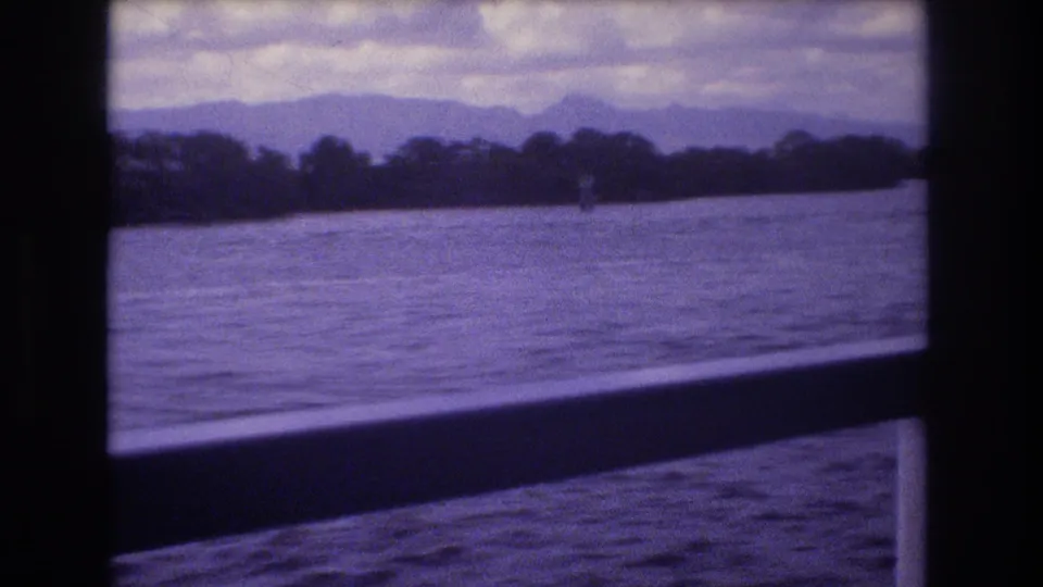 Handheld 8mm ferry footage from Honolulu showing rippled water, a low wooded shoreline, distant mountains, and a dark interior window bar.
