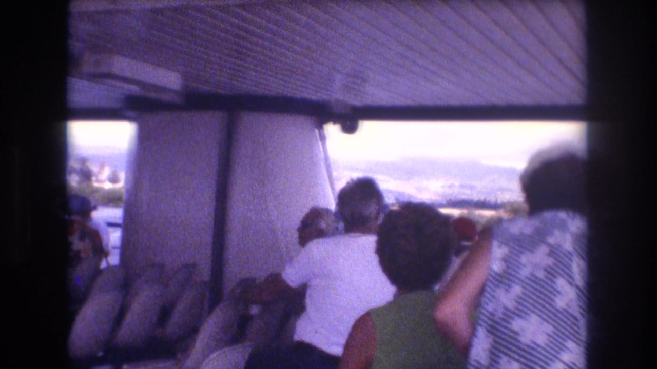 1977 Honolulu-area 8mm home-movie frame showing tourists seated under a covered boat canopy as they look out toward water and hazy mountains.