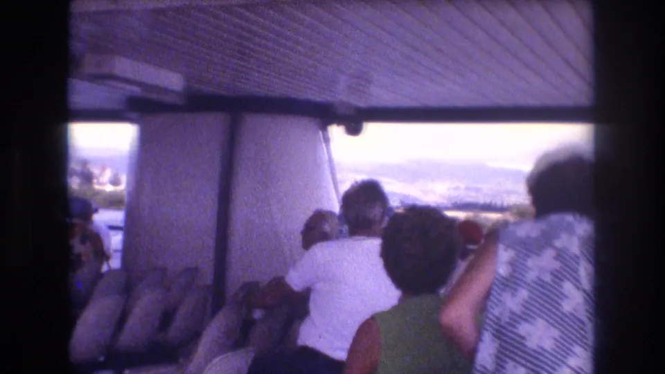 1977 Honolulu-area 8mm home-movie frame showing tourists seated under a covered boat canopy as they look out toward water and hazy mountains.