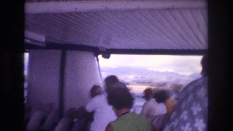 1977 Honolulu-area boat-tour frame with passengers in casual summer clothing facing outward toward water, shoreline, and distant hills.