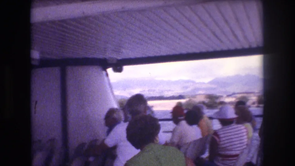 Soft 8mm frame from a Honolulu-area sightseeing boat, with tourists seated beneath the canopy and a bright water-and-mountain view outside.