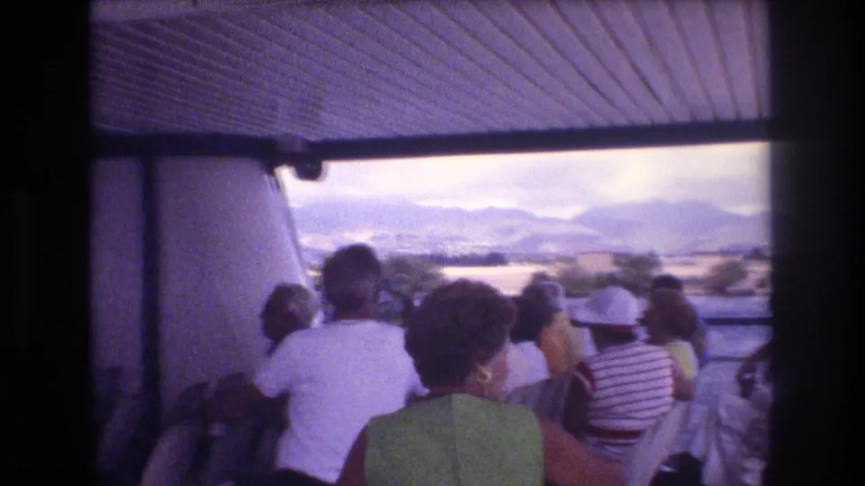 1977 Honolulu-area home-movie frame showing a covered tour boat interior with seated tourists watching the water, shoreline, and distant mountains.