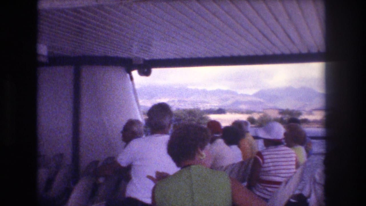 1977 Honolulu home-movie frame showing adult passengers on a covered boat tour facing open water, shoreline, and hazy mountains in the distance.