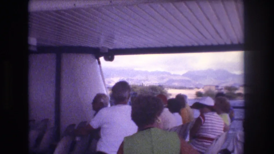 Adult tourists aboard a covered boat in Honolulu face away from the camera toward water and distant mountains, with casual summer clothing visible.