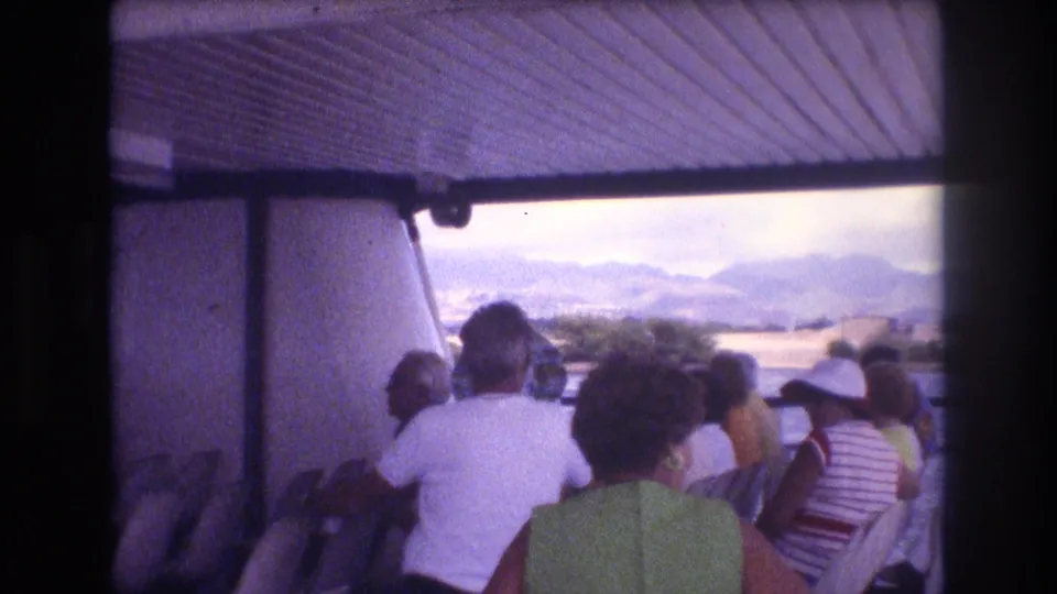 A steady 8mm frame shows Honolulu boat-tour passengers seated beneath a roof, looking past the vessel side toward water and mountainous shoreline.