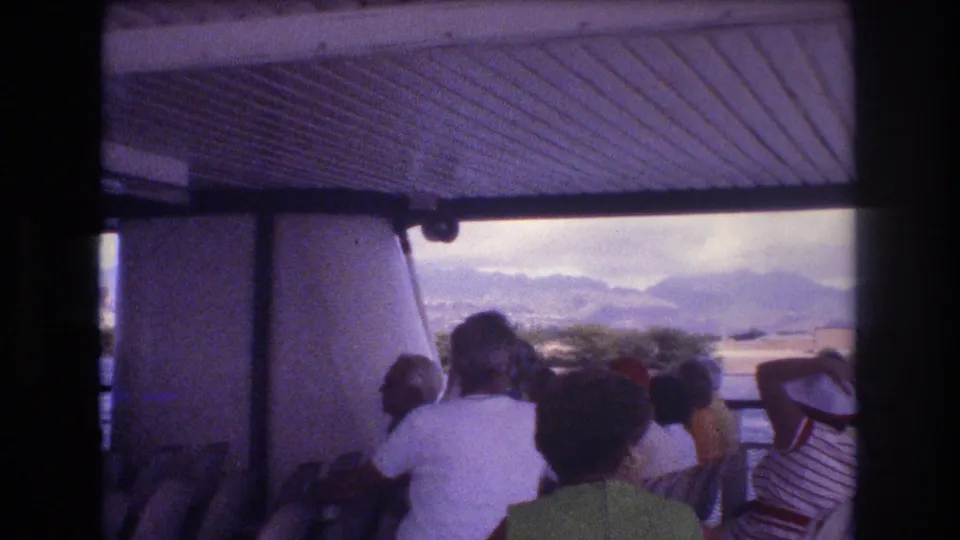 Late frame from the 1977 Honolulu boat-tour clip showing adults seated under a roof, one person raising an arm, with water and distant mountains beyond.