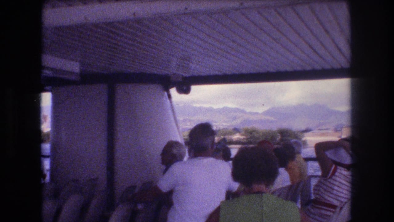 1977 Honolulu home-movie frame showing seated ferry passengers under a covered deck, facing the water and hazy mountains beyond.
