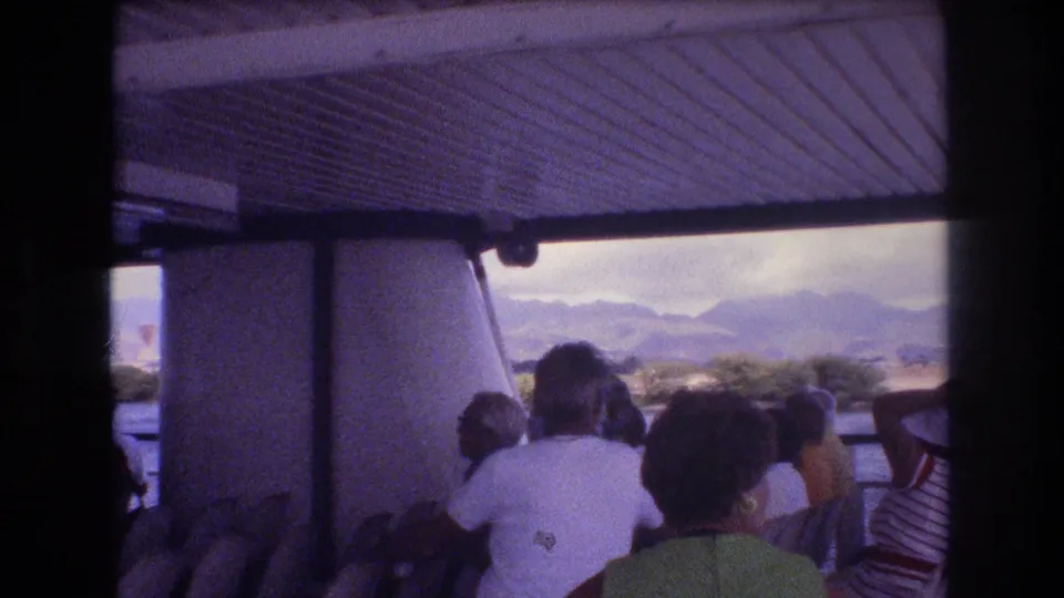 1977 Honolulu 8mm frame of tourists seated on an open-sided ferry, with water, shoreline, and distant hills visible through the deck opening.