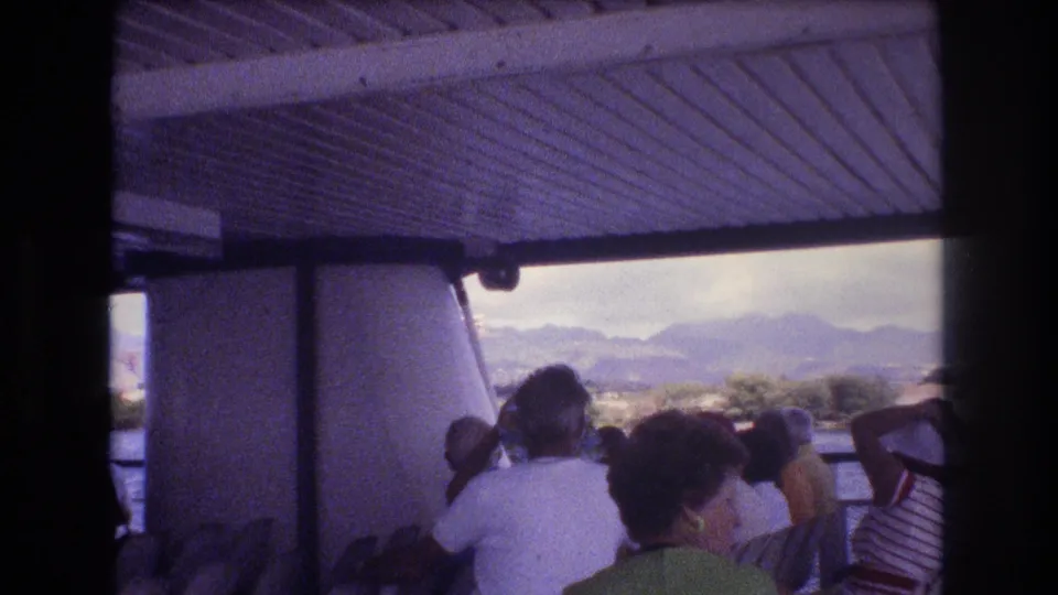 1977 Honolulu home-movie frame showing passengers on a covered ferry deck looking toward the water and mountains under a cloudy sky.