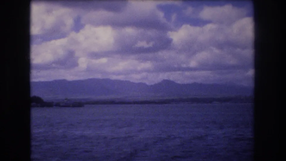 1977 Honolulu 8mm frame from a ferry showing open water, a low distant shoreline, mountains, and heavy clouds with no close passengers in view.