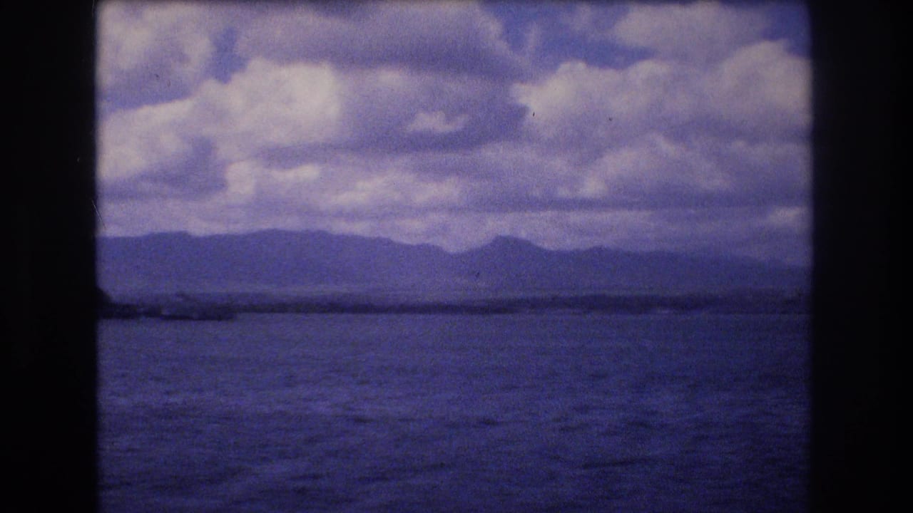 1977 Honolulu-area 8mm frame showing open water, a low distant shoreline or landform, mountain silhouettes, and heavy clouds through a dark film gate.