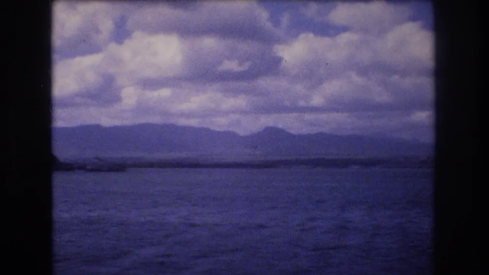 1977 Honolulu-area 8mm frame showing open water, a low distant shoreline or landform, mountain silhouettes, and heavy clouds through a dark film gate.