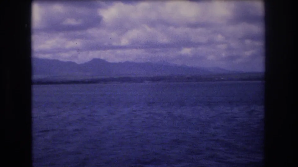1977 Honolulu-area boat-view frame with broad ocean foreground, hazy mountain ridge on the horizon, layered cloud cover, and visible 8mm frame edging.