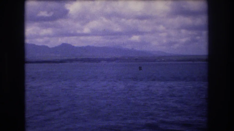 1977 Honolulu-area 8mm frame showing rippled ocean, a thin line of distant land, dark mountain shapes, and cloudy sky with dust and grain visible in the scan.