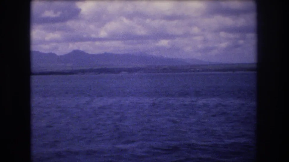 1977 Honolulu-area boat-view frame with textured ocean water, low coastline or land strip, distant mountains, dense clouds, and a pronounced purple-blue 8mm color cast.