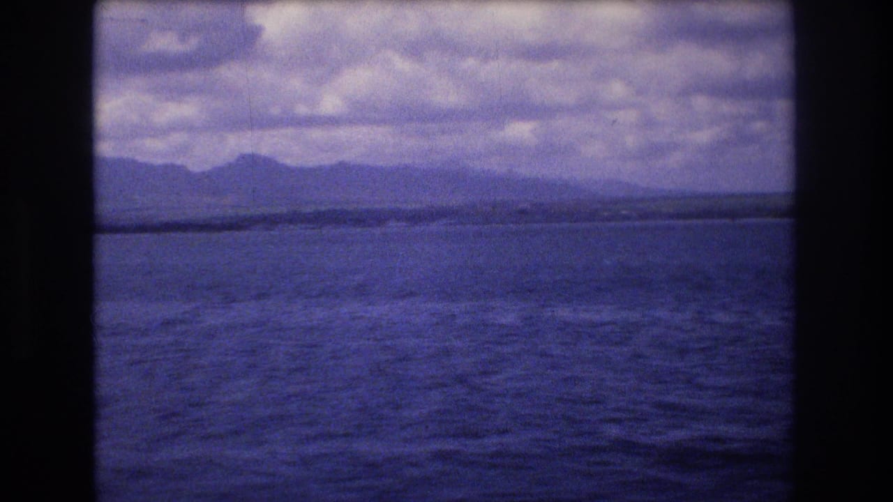 1977 Honolulu-area 8mm home-movie frame showing blue water, a low distant shoreline or island, and a cloudy sky with dark frame borders.