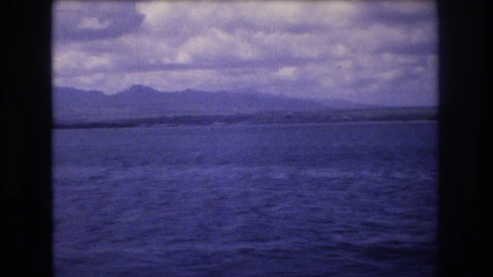 1977 Honolulu-area 8mm home-movie frame showing blue water, a low distant shoreline or island, and a cloudy sky with dark frame borders.