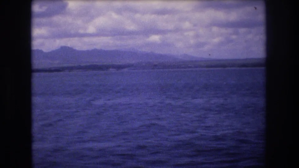 1977 Honolulu-area coastal panorama with calm water in the foreground, a distant hilly landform, and layered clouds across the sky.