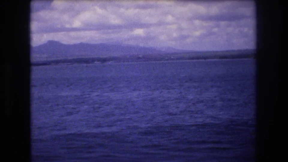 1977 Honolulu-area 8mm frame showing a broad expanse of water, a distant island or shoreline silhouette, and mottled cloud cover.